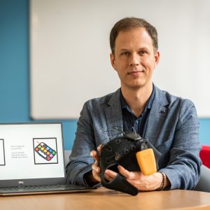 George Stothart is sat to a table. He is wearing a navy jacket and navy shirt. In his had is a black cap with wires attached. Next to him is a laptop with a photo of a pear on the left and a photo of a children's paint box on the right.