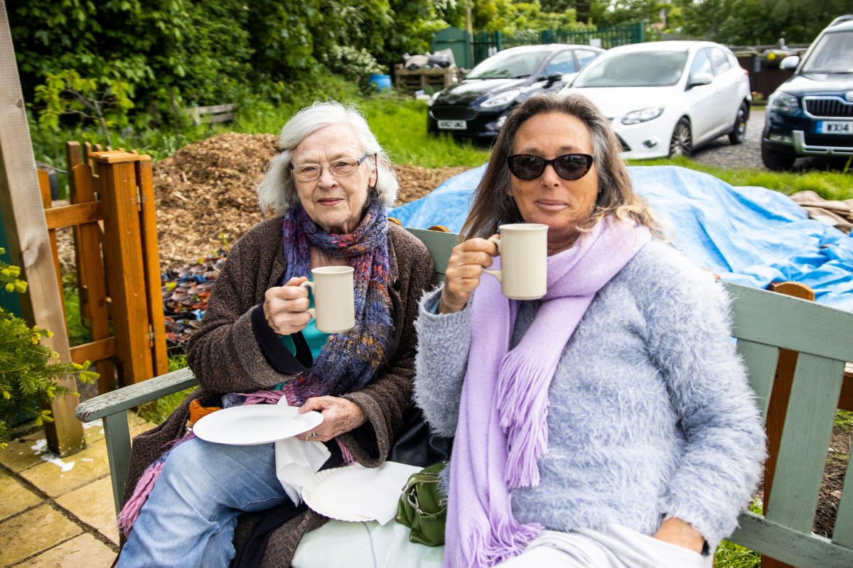 Two ladies are sat on a bench outside, with mugs of tea.