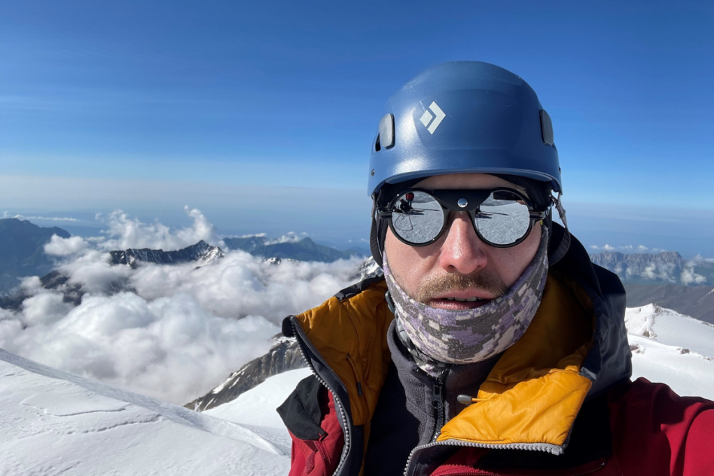 A man wearing on top of a snowy mountain, wearing a helmet, jacket and reflective sunglasses.