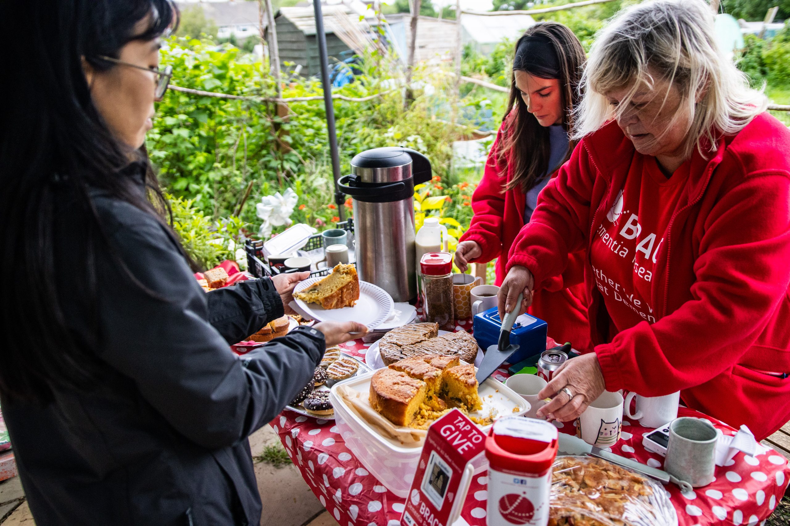 Two women in red fleeces are serving cake and hot drinks to another woman.