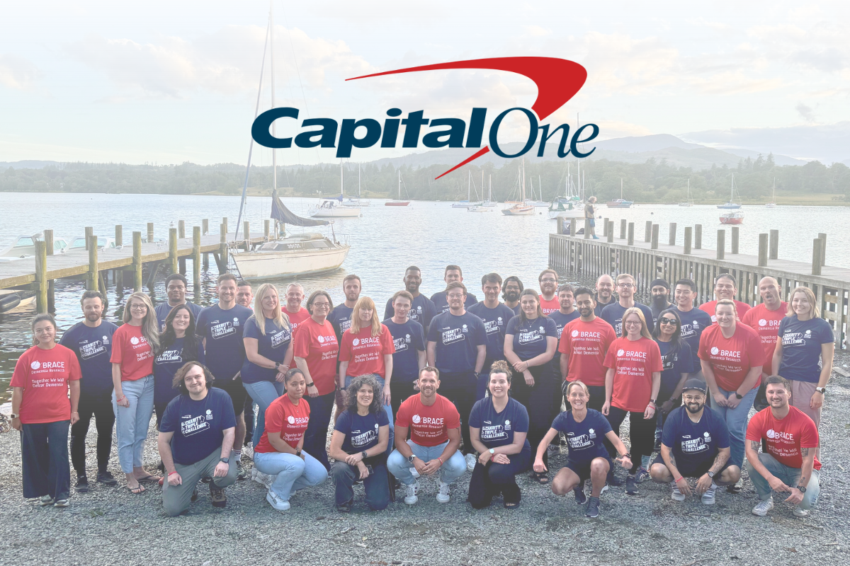 A large group of people wearing BRACE charity t-shirts in red and navy t-shirts pose by a lakeside with wooden docks and sailboats in the background. The 'Capital One' logo is displayed above them.