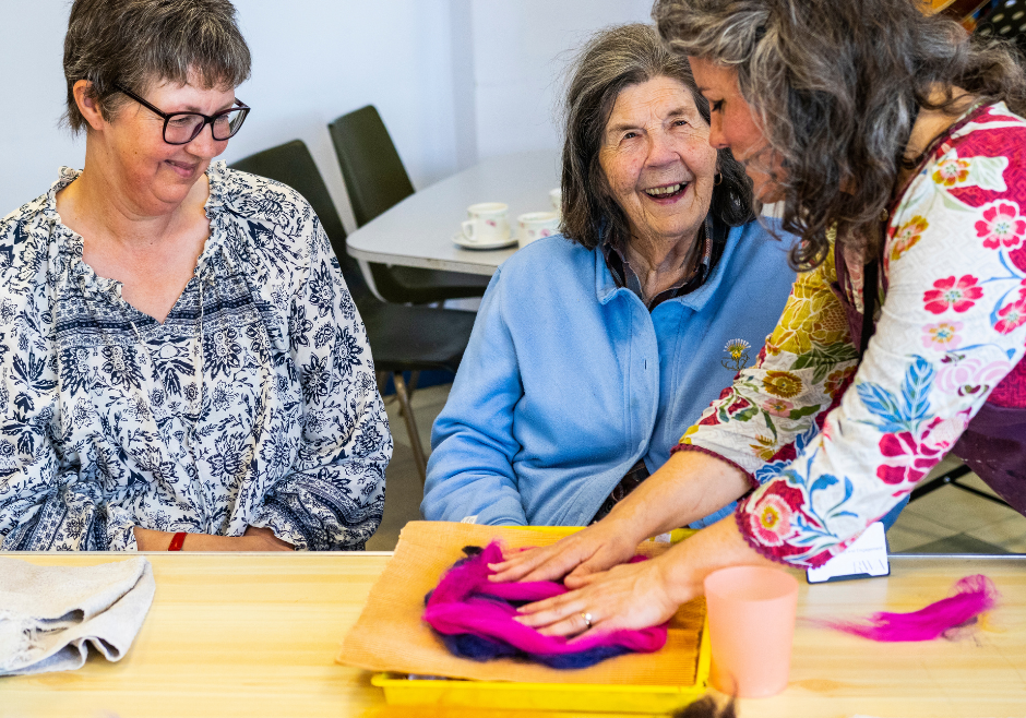 A woman sat at a table smiles as she is taught how to make felt art.