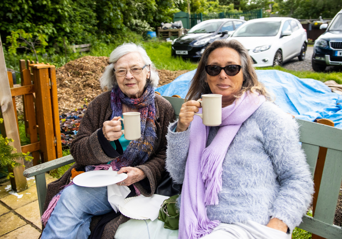 Two women are sat on a bench drinking tea and holding plates with cake on.